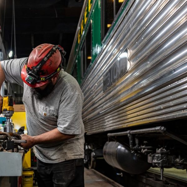 Worker in red helmet using a power tool next to a shiny metal train in a workshop.