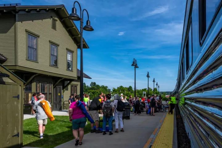 People standing on a train station platform beside a building and train under a clear blue sky.
