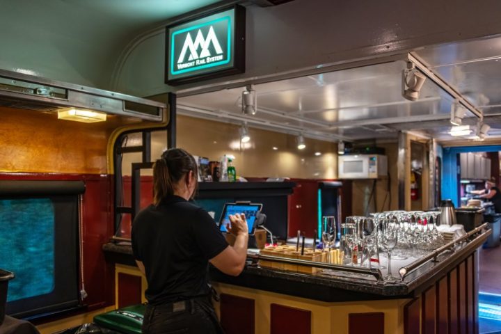 Person using touchscreen at train bar with glasses and Vermont Rail System sign.