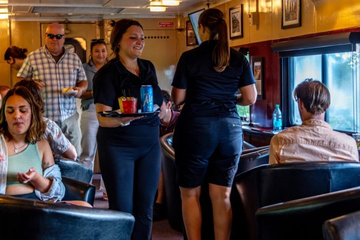 Train car interior with people, including servers carrying trays amidst seated passengers.