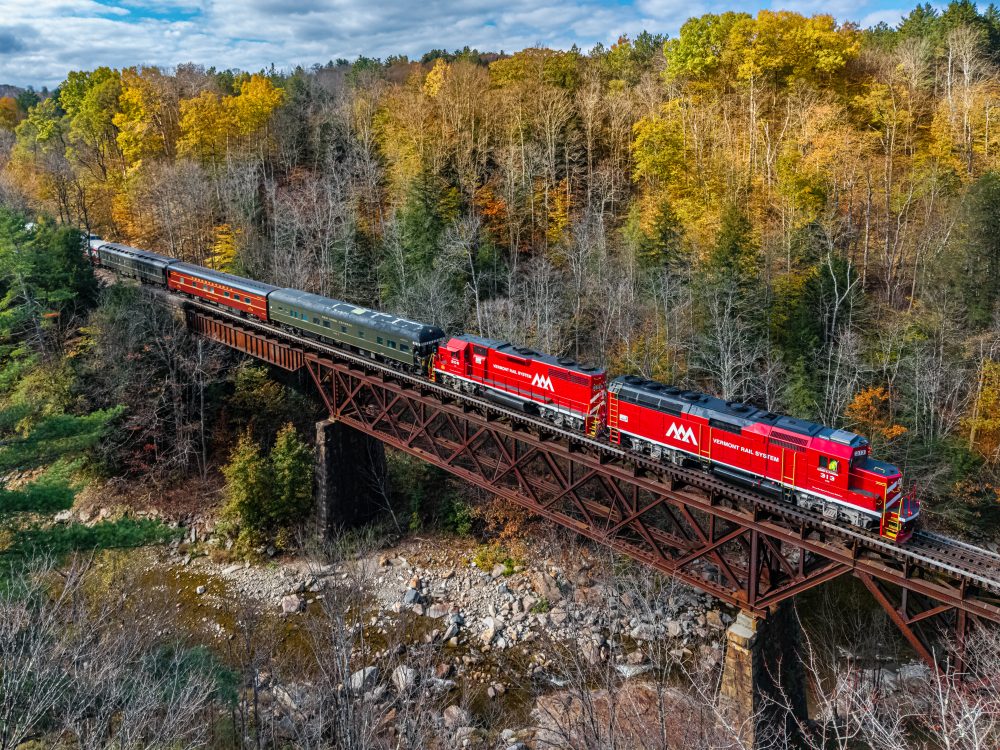 Red train crossing a trestle bridge in a forest with autumn foliage.
