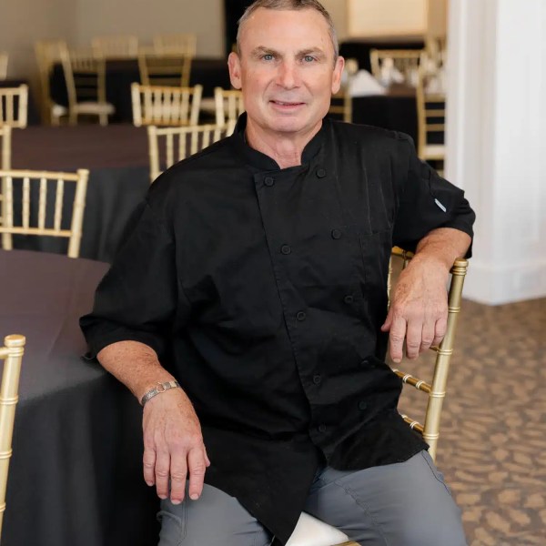 Chef in black uniform sitting on a chair in a dining area with gold chairs.