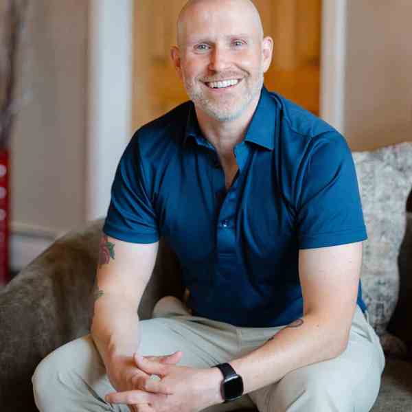 Smiling bald man in blue shirt and beige pants seated on a sofa.