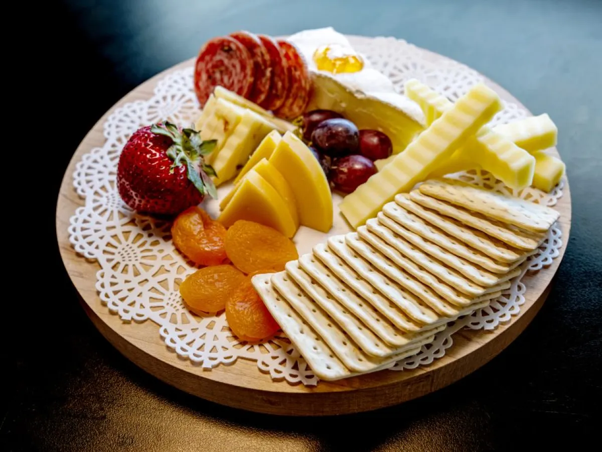 Cheese board with crackers, sliced meats, cheeses, dried apricots, strawberries, and grapes on a doily.