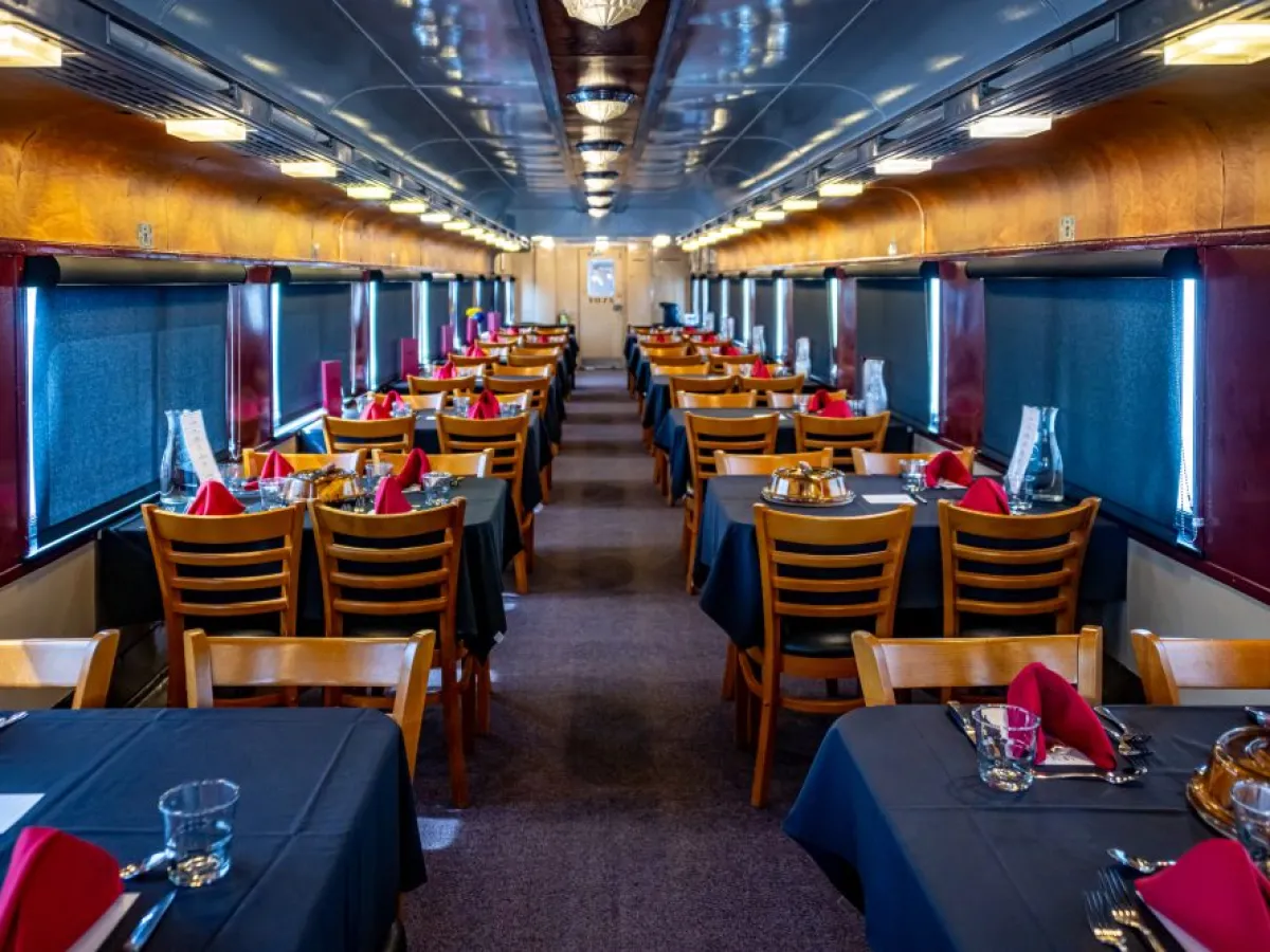Train dining car with wooden chairs and tables set for meal, red napkins and black tablecloths.