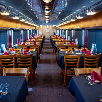Train dining car with wooden chairs and tables set for meal, red napkins and black tablecloths.