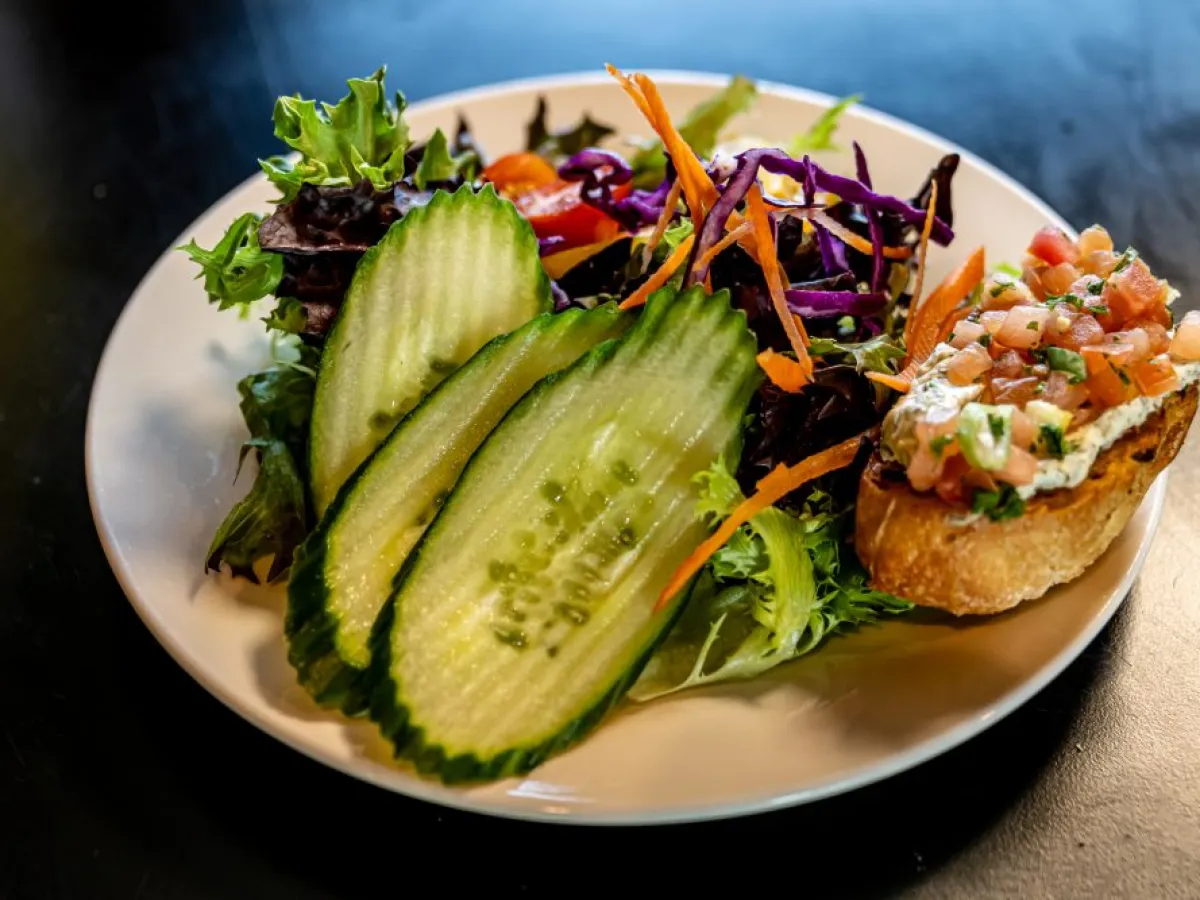 Plate with mixed salad, cucumber slices, and bruschetta topped with tomato on a dark surface.