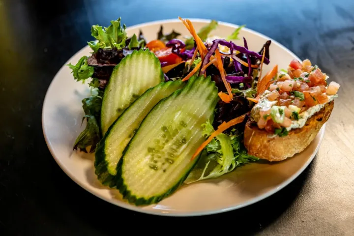Plate with mixed salad, cucumber slices, and bruschetta topped with tomato on a dark surface.