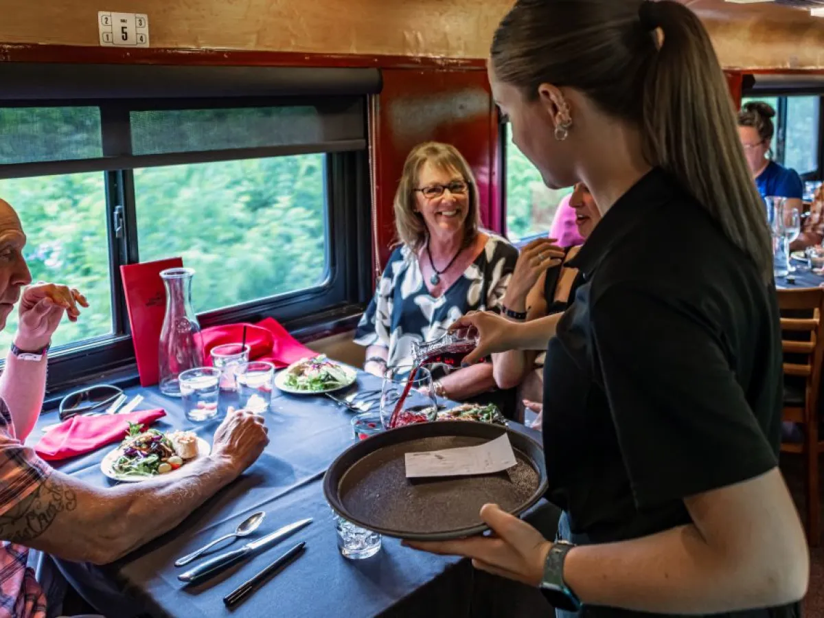 Server pouring wine for smiling diners on a train, with plates of food on a table near a window.