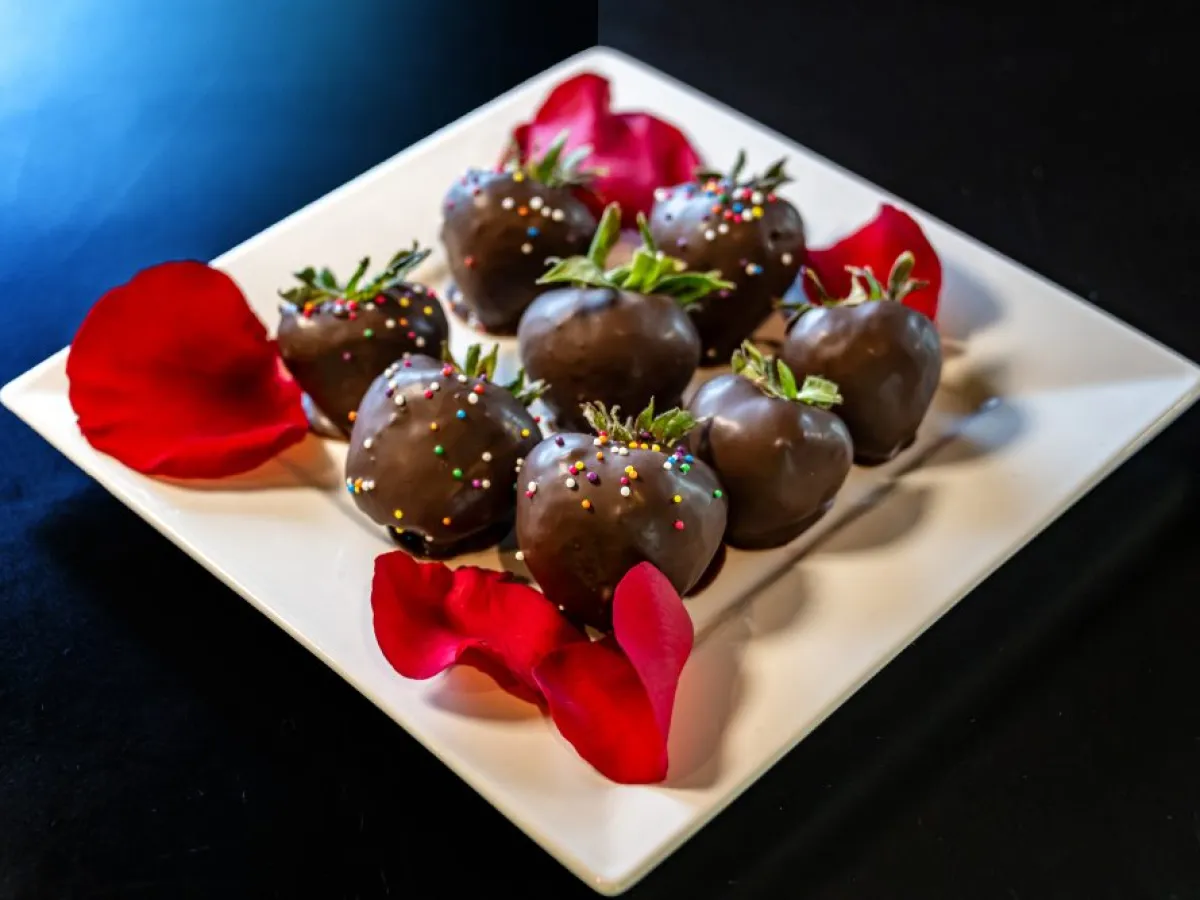 Chocolate-covered strawberries with sprinkles on a white plate, surrounded by red rose petals.
