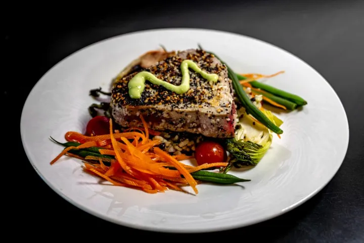 Seared tuna steak with sesame seeds, topped with sauce, served with vegetables on a white plate.