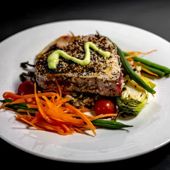 Seared tuna steak with sesame seeds, topped with sauce, served with vegetables on a white plate.