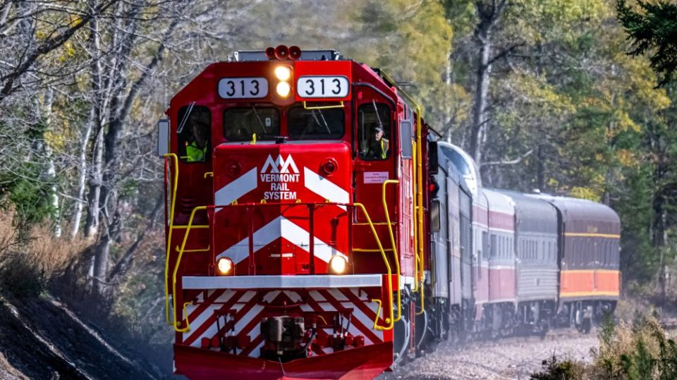 Red train with Vermont Rail System logo travels through forested area.