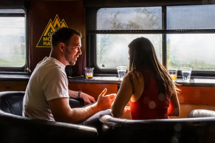 Man and woman talking in a train with drinks on a table, sunlight streaming through windows.