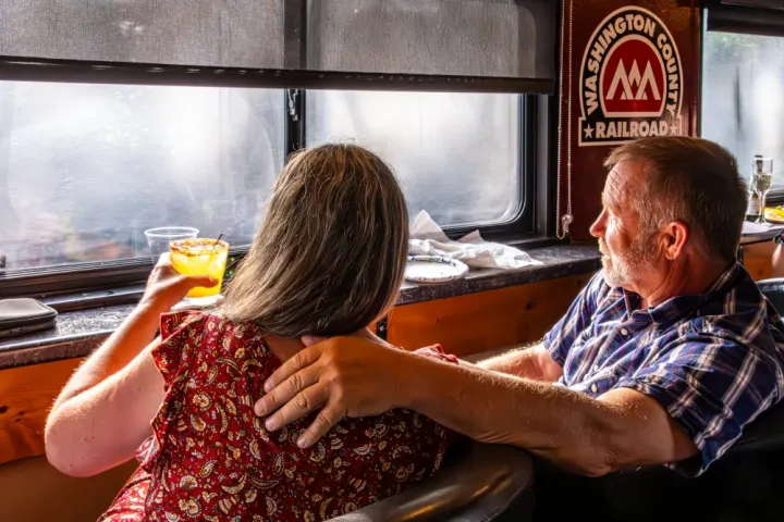 Couple on a train, woman holds a drink, man's arm around her, 'Washington County Railroad' sign visible.