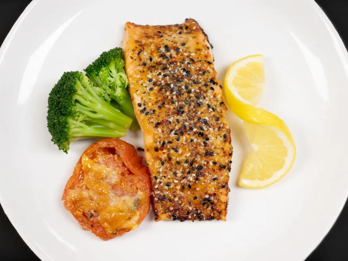 Grilled salmon fillet with broccoli, baked tomato, and lemon slices on a white plate.