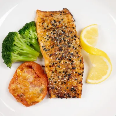 Grilled salmon fillet with broccoli, baked tomato, and lemon slices on a white plate.