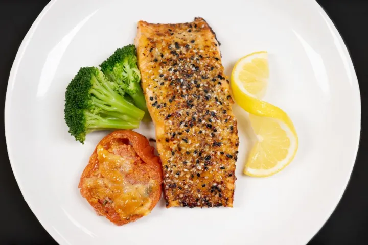 Grilled salmon fillet with broccoli, baked tomato, and lemon slices on a white plate.