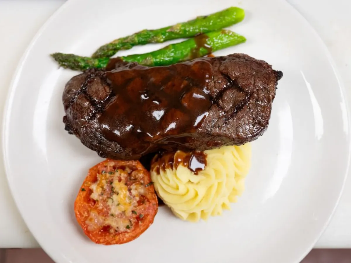 Grilled steak with sauce, mashed potatoes, tomato, and asparagus on a white plate.