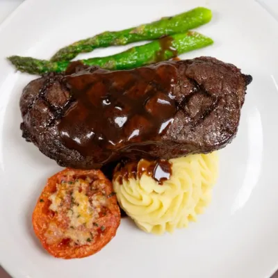 Grilled steak with sauce, mashed potatoes, tomato, and asparagus on a white plate.