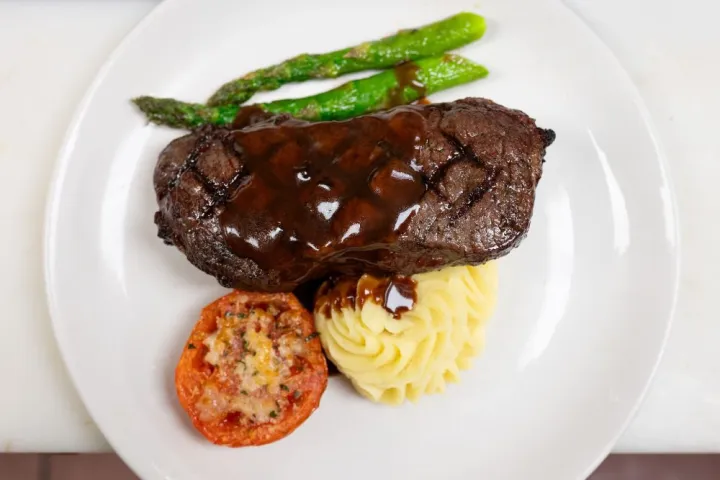 Grilled steak with sauce, mashed potatoes, tomato, and asparagus on a white plate.